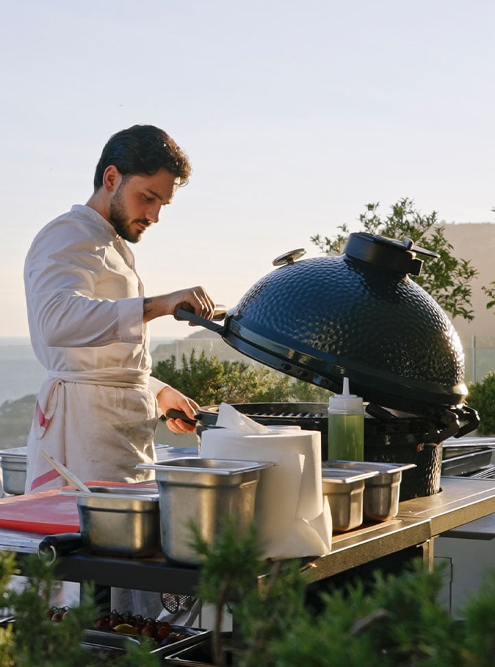 A chef in a white uniform lifts the lid of a large outdoor ceramic grill, preparing food at sunset with ocean views.